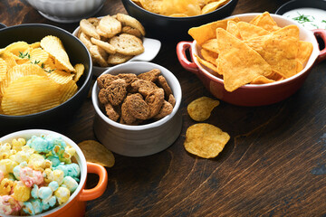 Unhealthy food or snacks. All classic potato snacks with peanuts, popcorn and onion rings and salted pretzels in bowl plates on old wooden background. Unhealthy food for figure, heart, skin, teeth.