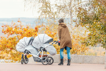 Mother woman walks with twin baby stroller or pushchair in autumn park