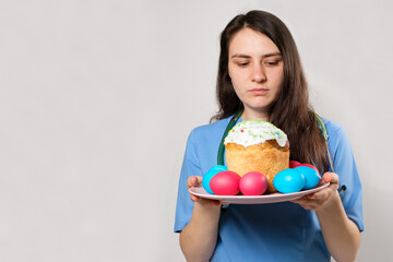 A medical worker in blue uniform holds an Easter cake and painted eggs on a white background, a place for text.