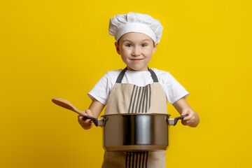 Isolated picture of a happy Caucasian cheerful male child in a chef's hat and apron, holding a saucepan in his hands, smiling at the camera on a yellow background
