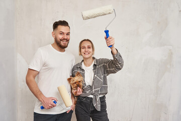 Renovation diy paint couple in new home painting wall together. cheerful young couple and a small dog on the background of repair with paint rollers in their hands