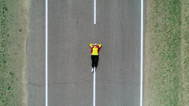 Aerial top down view of a young woman with pink hair braids and yellow jacket laying on an asphalt road.