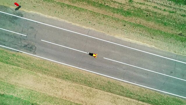 Aerial top down view of a young woman with pink hair braids and yellow jacket laying on an asphalt road.