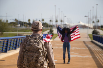 African American woman with American flag, waiting for her partner, an American soldier who has just arrived from a mission. Concept army, missions, military, war. Selective focus on the soldier.