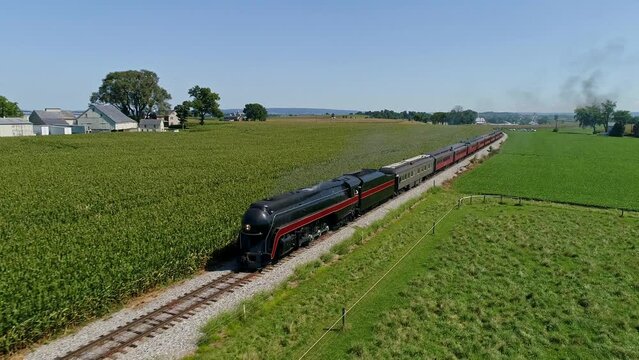 An Aerial View Traveling In Front of An Antique Steam Passenger Train, Blowing Smoke Traveling Thru Corn Fields and Rich Farmlands on a Cloudless Summer Day