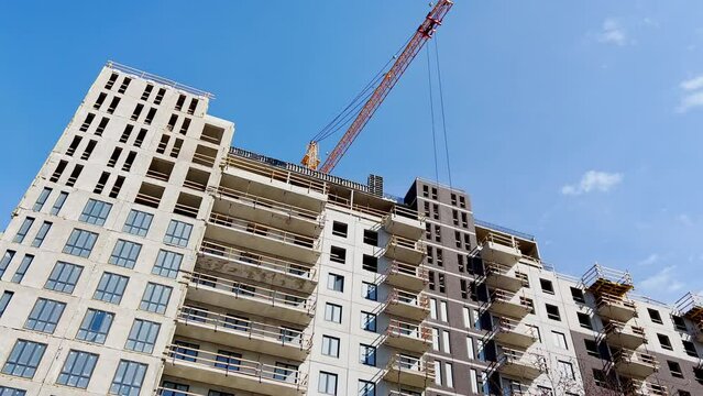 Construction of the high floor house at sunny day, the worker in a protective helmet does a facade, construction cranes, the bottom view up, concrete blocks with windows