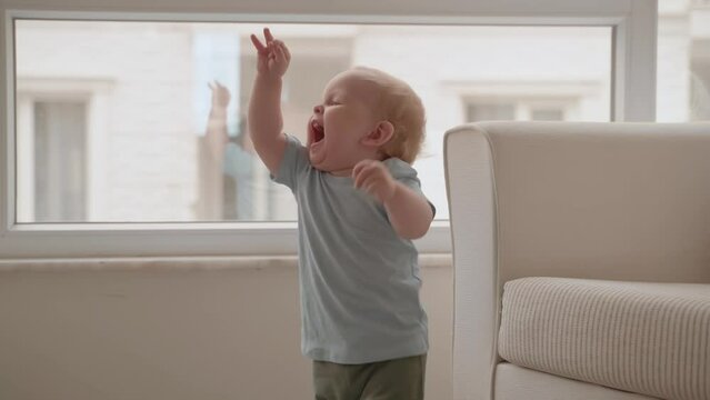Cheerful baby boy trying to do his first steps in living room, getting hew skill happy. Curious laughing infant enjoying his happy childhood. Mother teaching her toddler son how to walk. Slow Motion