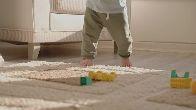 Little baby boy taking his first steps without help on floor in light living room. Close up of bare tiny feet doing first steps on camera. Toddler child learning to walk. Childhood. Slow motion