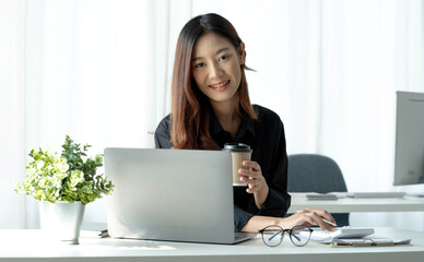 Smiling Asian businesswoman holding a coffee mug and laptop at the office. Looking at the camera.