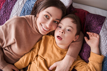 Boy with Down syndrome laying at the bed with his mother at the morning