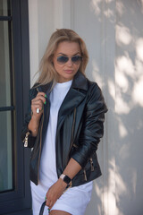 teen style. young girl in white oversize dress and black leather jacket stands casual in sunglasses and smiles cute on a white street cafe background at sunny day. sport lifestyle concept, free space