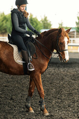 Obraz premium Young girl in helmet and special clothes riding a horse on the sandy ground of the paddock
