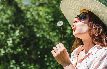 Young happy curly woman wearing hat and sunglasses blowing on a dandelion in nature.