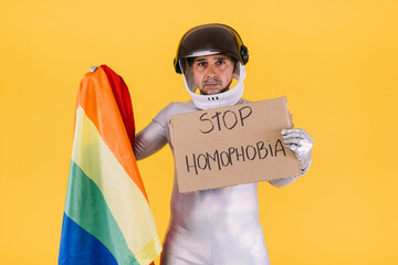 Gay man dressed as an astronaut with a helmet and silver suit, with the flag of the lgtbi collective holding a sign that reads: 'stop homophobia', on a yellow background.
