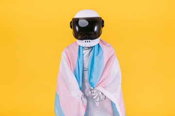 Gay man dressed as an astronaut with a helmet and silver suit, holding a flag of the trans community, on a yellow background. Gay, homosexual, trans, rights and gender pride concept.