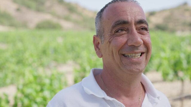 Happy Caucasian male winemaker smiling looking away with blurred plantation at background. Close-up portrait of satisfied confident man admiring field on Cyprus. Cultivation and horticulture