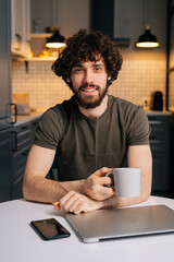 Vertical portrait of successful bearded young freelancer male holding in hand cup with morning coffee sitting at table with closed laptop computer, looking at camera, in kitchen with modern interior.