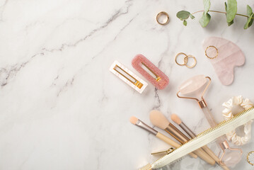 Fototapeta na wymiar Top view photo of pink hairpins rose quartz roller gua sha golden rings transparent cosmetics bag with makeup brushes scrunchy and eucalyptus on white marble background with blank space