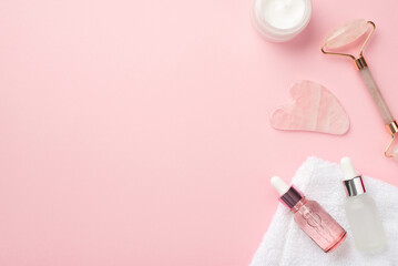 Obraz na płótnie Canvas Skincare concept. Top view photo of rose quartz roller gua sha massager cream jar two glass transparent bottles and white towel on pastel pink background with blank space