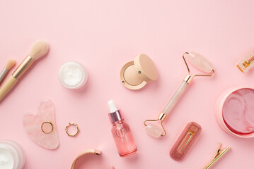 Top view photo of makeup brushes rose quartz roller gua sha pink eye patches glass transparent bottle cream jars eyeshadow barrettes lip gloss gold rings and wristlet on isolated pink background