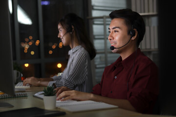 Happy smiling Asian man work at call center service desk consultant with teammates at night, customer service executive with microphone headset use computer for supporting in late night office.