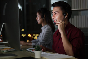 Happy smiling Asian man work at call center service desk consultant with teammates at night, customer service executive with microphone headset use computer for supporting in late night office.