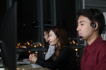 Happy smiling Asian man work at call center service desk consultant with teammates at night, customer service executive with microphone headset use computer for supporting in late night office.