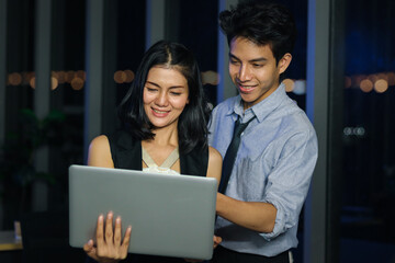 Happy smiling young Asian man and woman teammate officer have overwork project overnight in office, use laptop computer for discussing job at mid night, work  hard overload night-shift business people