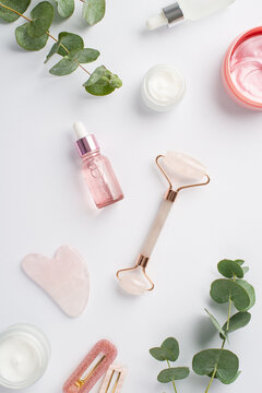 Skincare concept. Top view vertical photo of rose quartz roller gua sha pink eye patches glass bottles barrettes and eucalyptus on isolated white background
