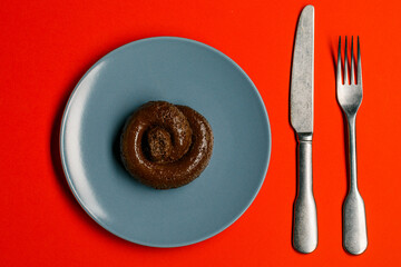 Plastic excrement lies on a plate between a fork and a knife close-up. The concept of an inept cook, fake news, shitty restaurant, bad food, taste of shit