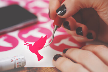 women hands with painted nails hold freshly cut peace dove decal that sticks to tip of curved weeding tool. pink adhesive film. wooden worktop. adjustable plotting blade in foreground. selective focus - obrazy, fototapety, plakaty