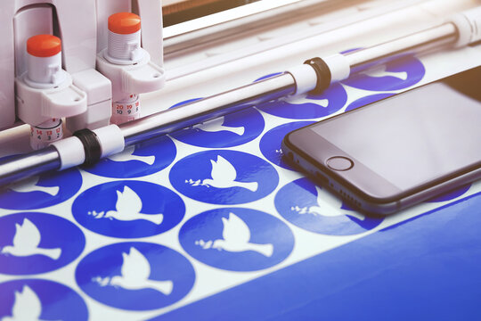 multiple round peace dove stickers are made by a plotting machine from blue adhesive film. view on tool head, guides, uncut film and mobile phone. selctive focus. warm light