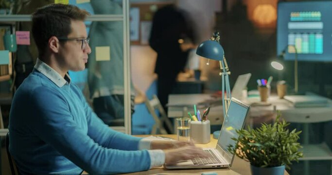 Zoom-in time lapse of successful entrepreneur working with laptop and collaborating with colleagues in office at night. Job and technology concept.
