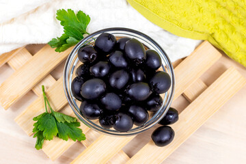 Many black marinated whole olives in the glass bowl on light wooden board background in the kitchen top view.