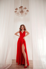 Fototapeta premium teen fashion. young blonde girl in long red dress is standing with hands near waist and looking straight with smile on the white wall background with crystal chandelier. fashion concept, free space
