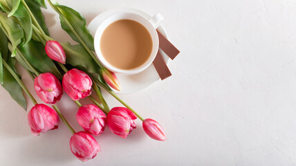 Banner.One cup of hot morning coffee, milk chocolate and a bouquet of pink-lilac tulips on a bright white background. View from above. Copy space for text. Holidays concept.