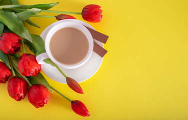 Banner.One cup of hot morning coffee, milk chocolate and a bouquet of pink-lilac tulips on a bright yellow background. View from above. Copy space for text. Holidays concept.