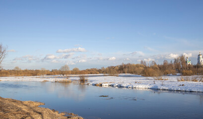 Fototapeta premium March sunny day by the river. A picturesque landscape, early spring, a river with snow-covered banks, dry grass and bushes. Church in the background. The first thaws, the snow is melting