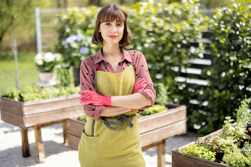 Portrait of young and cheerful gardener in green apron standing at home vegetable garden in...