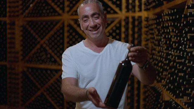 Proud confident winemaker stretching bottle looking at camera standing in wine cellar in darkness. Portrait of satisfied Caucasian man posing in bodega indoors in slow motion. Quality and confidence