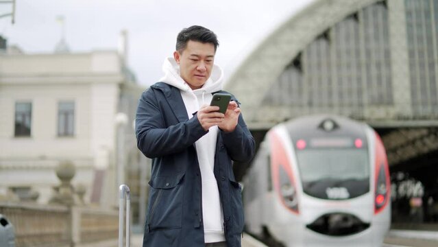 Asian tourist booking accommodation at home using mobile phone, passenger arrives by train to new city. Passengers at rairoad station and arriving train. Man use smart phone before travel
