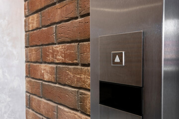 Button to call the elevator up on a metal panel with a red brick wall
