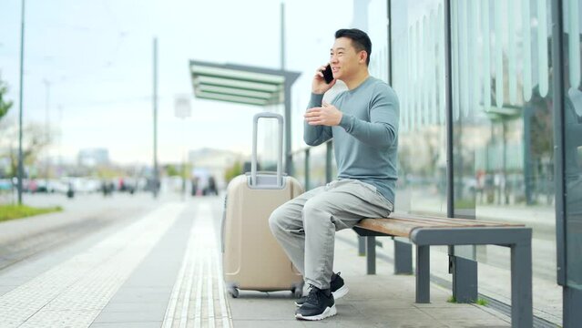 Happy Chinese tourist talk mobile phone app to book accommodation and call taxi, man near airport with suitcase. Asian man conversation to rent a car at the bus stop. waiting public transport
