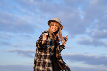 Young caucasian girl holding smartphone and credit card happy, crazy or amazed for success. Excited hipster tourist woman wearing shirt and hat makes shocked winner gesture outdoor with sky background