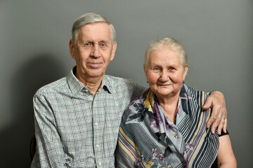 Studio portrait of an elderly couple on a gray background. The old man is hugging his wife, they are smiling, happy.