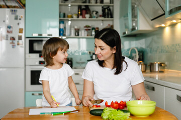 Happy smiling family mom and daughter together in the kitchen. The girl draws while her mother makes a salad of fresh vegetables.