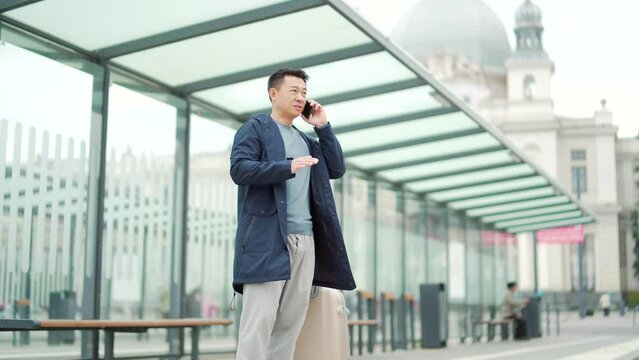 Happy Chinese tourist talk mobile phone app to book accommodation and call taxi, man near airport with suitcase. Asian man conversation to rent a car at the bus stop. waiting public transport