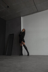 Fototapeta na wymiar fashion leisure. young teen girl in black dress-jacket and over the knee boots stands casual and looks apart with cute smile near gray studio wall background with mirror. lifestyle concept, free space