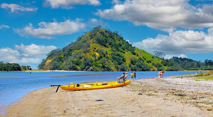 Pataua, New Zealand - December 9. 2012: Beautiful sand beach with canoe, people relaxing in sun, water bay, lush green hill, blue sky fluffy cumulus clouds