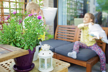 Obraz na płótnie Canvas Happy kids have fun and pillow fight on cozy balcony at home. Wooden table, many green plants, soft sofa, candles. Lovely terrace life on vacation. Green place in city apartment. Focus on flower